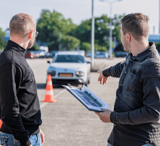 Two men speaking about a car, one holding a clipboard pointing at the car with cones around it
