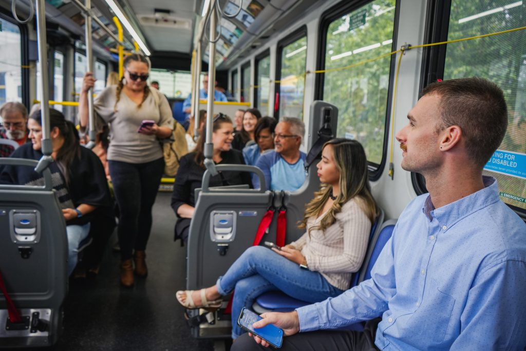 Many people on bus sitting with a woman standing holding hand rail